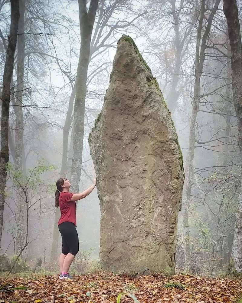 une femme face au menhir imposant du Jardin de Merlin, au milieu de la forêt de Rosheim,