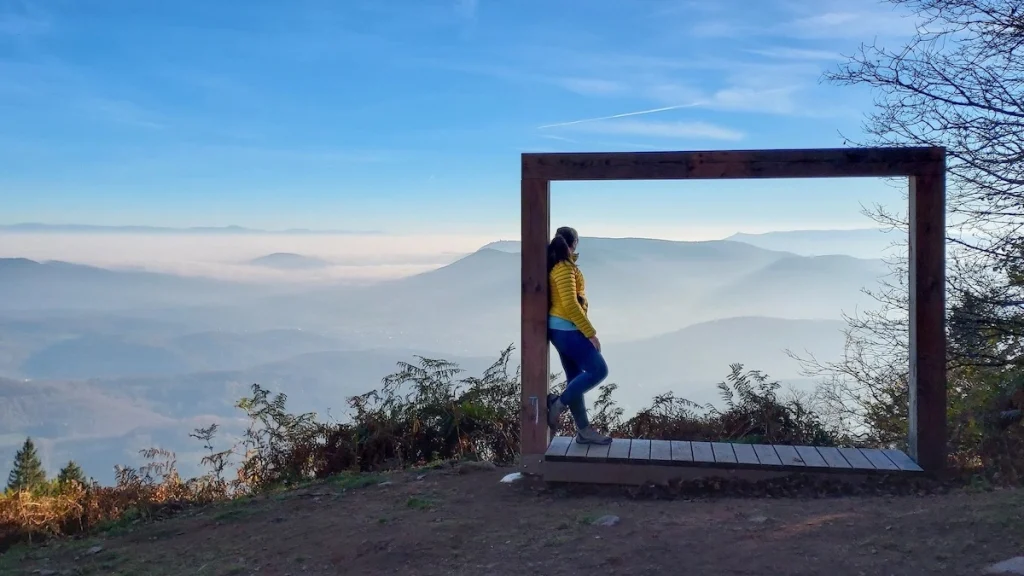 femme randonneuse en doudoune jaune qui prend pause photo dans un grand cadre en bois avec au fond, la ligne des crêtes vosgiennes